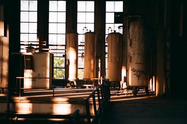 A dimly lit industrial space with large cylindrical tanks and machinery inside an old building. The sunlight streaming through tall, dusty windows casts long shadows, creating a dramatic play of light and shadow. Some graffiti can be seen on one of the tanks, adding a touch of urban decay.