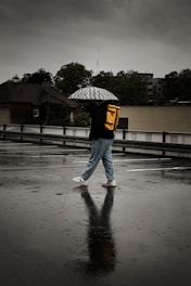 A person walks across a wet, reflective surface while holding an umbrella with a patterned design. They wear a yellow backpack, denim jeans, and sneakers. The background includes overcast skies and trees, set against a backdrop of urban buildings with gloomy, dark clouds overhead.