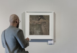 An individual with glasses and a beard observes a framed painting hung on a wall. The artwork features a nude form with muted colors and minimalistic detail. The observer is dressed in a gray jacket and carries a bag over one shoulder, gazing intently at the art.