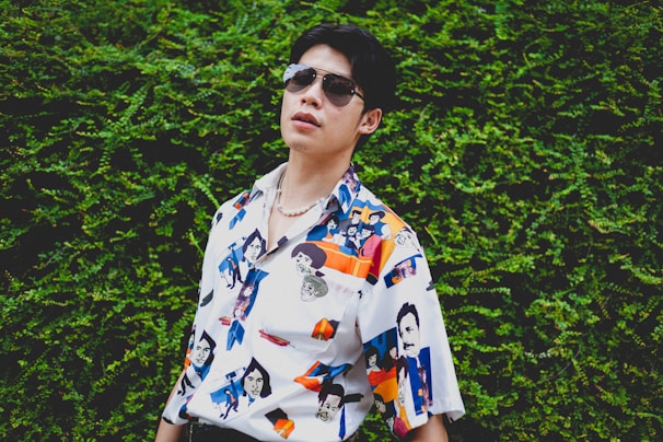 A person wearing sunglasses and a colorful, patterned shirt featuring various illustrations and faces stands against a lush green leafy background. The shirt's design includes abstract and pop-art elements, enhancing its vibrant aesthetic.
