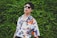 A person wearing sunglasses and a colorful, patterned shirt featuring various illustrations and faces stands against a lush green leafy background. The shirt's design includes abstract and pop-art elements, enhancing its vibrant aesthetic.