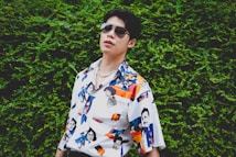A person wearing sunglasses and a colorful, patterned shirt featuring various illustrations and faces stands against a lush green leafy background. The shirt's design includes abstract and pop-art elements, enhancing its vibrant aesthetic.