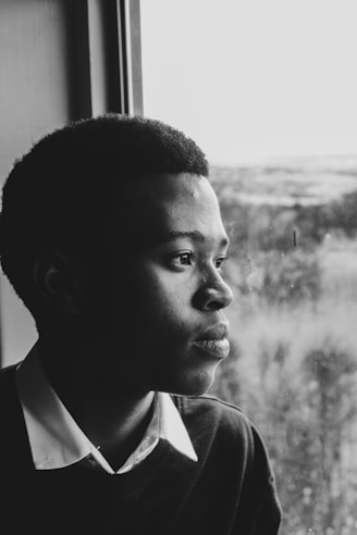 A black and white portrait of a woman looking thoughtfully out of a window with soft natural light.