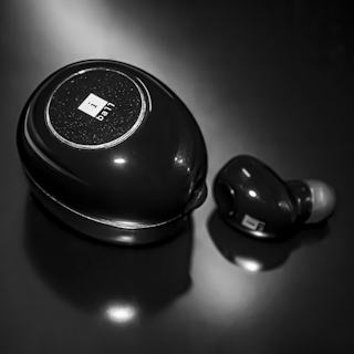 A close-up of a black earbud and its charging case, both featuring a glossy finish and a circular design. The case has a shiny emblem or logo on its surface, and the earbud has a small rubber tip.