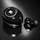 A close-up of a black earbud and its charging case, both featuring a glossy finish and a circular design. The case has a shiny emblem or logo on its surface, and the earbud has a small rubber tip.