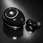A close-up of a black earbud and its charging case, both featuring a glossy finish and a circular design. The case has a shiny emblem or logo on its surface, and the earbud has a small rubber tip.