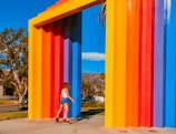 A person on roller skates moves near a large, colorful geometric structure composed of vertical, rainbow-colored panels. The sky is clear blue, and trees are visible in the background, suggesting a sunny day in an outdoor setting.