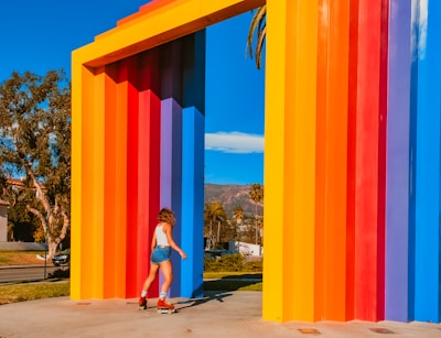 A person on roller skates moves near a large, colorful geometric structure composed of vertical, rainbow-colored panels. The sky is clear blue, and trees are visible in the background, suggesting a sunny day in an outdoor setting.