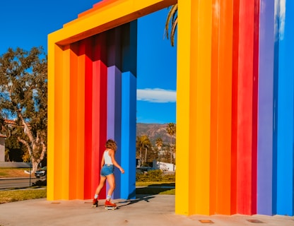 A person on roller skates moves near a large, colorful geometric structure composed of vertical, rainbow-colored panels. The sky is clear blue, and trees are visible in the background, suggesting a sunny day in an outdoor setting.