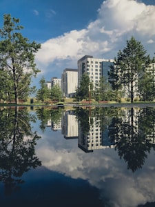 High-rise apartment buildings are reflected in a perfectly still body of water, creating a mirror image effect. The setting is enhanced by lush green trees and a clear blue sky filled with white clouds.