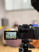 A digital camera is positioned on a tripod, with its LCD screen displaying a scene being recorded. The background appears blurred, suggesting a depth of field focusing on the camera.