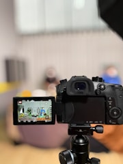 A digital camera is positioned on a tripod, with its LCD screen displaying a scene being recorded. The background appears blurred, suggesting a depth of field focusing on the camera.