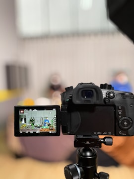 A digital camera is positioned on a tripod, with its LCD screen displaying a scene being recorded. The background appears blurred, suggesting a depth of field focusing on the camera.