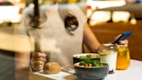 A person is sitting at a table with a meal consisting of a bowl of broccoli, a bread roll, a yogurt parfait, and a glass of orange juice with mint leaves. The person is holding a smartphone in their right hand.