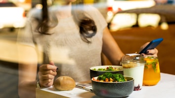 A person is sitting at a table with a meal consisting of a bowl of broccoli, a bread roll, a yogurt parfait, and a glass of orange juice with mint leaves. The person is holding a smartphone in their right hand.
