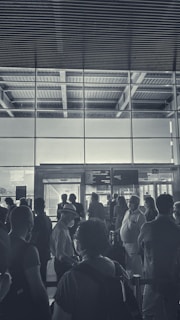 A group of people is gathered in an airport terminal near a boarding gate. The scene includes individuals of various ages, some wearing masks and hats, standing in a queue or waiting area. The architecture features large glass windows allowing light to stream through, highlighting the industrial ceiling design above.