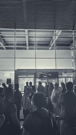 A group of people is gathered in an airport terminal near a boarding gate. The scene includes individuals of various ages, some wearing masks and hats, standing in a queue or waiting area. The architecture features large glass windows allowing light to stream through, highlighting the industrial ceiling design above.