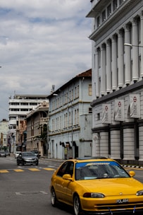 A street scene featuring a vibrant yellow car in the foreground, driving along a road lined with historical buildings. The architecture includes ornate and colonial style elements, with a prominent building displaying HSBC signs. The sky is cloudy, indicating an overcast day.