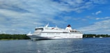 A large white cruise ship with a red and blue stripe moves gracefully across a calm body of water. The ship is detailed with multiple decks and the name 'Cinderella' is visible along the side. In the background, lush green trees line the shoreline under a partly cloudy, blue sky.