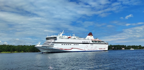 A large white cruise ship with a red and blue stripe moves gracefully across a calm body of water. The ship is detailed with multiple decks and the name 'Cinderella' is visible along the side. In the background, lush green trees line the shoreline under a partly cloudy, blue sky.