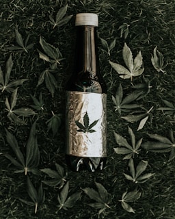 A dark-colored bottle wrapped in reflective foil is placed on grass, surrounded by scattered cannabis leaves. The foil features a printed cannabis leaf.