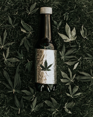 A dark-colored bottle wrapped in reflective foil is placed on grass, surrounded by scattered cannabis leaves. The foil features a printed cannabis leaf.
