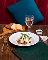 A tastefully arranged dish sits on a white plate, garnished with fresh herbs, likely rosemary. The dish is placed on a wooden table, accompanied by utensils, a folded green napkin, and a wooden board with slices of brown and white bread. A glass of white wine stands nearby, and small bowls of spices add a touch of culinary detail. A plush red seating area with a teal pillow provides a cozy background.