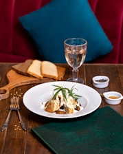 A tastefully arranged dish sits on a white plate, garnished with fresh herbs, likely rosemary. The dish is placed on a wooden table, accompanied by utensils, a folded green napkin, and a wooden board with slices of brown and white bread. A glass of white wine stands nearby, and small bowls of spices add a touch of culinary detail. A plush red seating area with a teal pillow provides a cozy background.