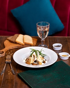 A tastefully arranged dish sits on a white plate, garnished with fresh herbs, likely rosemary. The dish is placed on a wooden table, accompanied by utensils, a folded green napkin, and a wooden board with slices of brown and white bread. A glass of white wine stands nearby, and small bowls of spices add a touch of culinary detail. A plush red seating area with a teal pillow provides a cozy background.