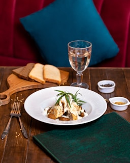 A tastefully arranged dish sits on a white plate, garnished with fresh herbs, likely rosemary. The dish is placed on a wooden table, accompanied by utensils, a folded green napkin, and a wooden board with slices of brown and white bread. A glass of white wine stands nearby, and small bowls of spices add a touch of culinary detail. A plush red seating area with a teal pillow provides a cozy background.