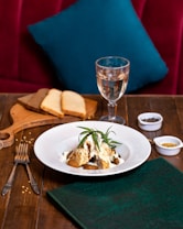 A tastefully arranged dish sits on a white plate, garnished with fresh herbs, likely rosemary. The dish is placed on a wooden table, accompanied by utensils, a folded green napkin, and a wooden board with slices of brown and white bread. A glass of white wine stands nearby, and small bowls of spices add a touch of culinary detail. A plush red seating area with a teal pillow provides a cozy background.