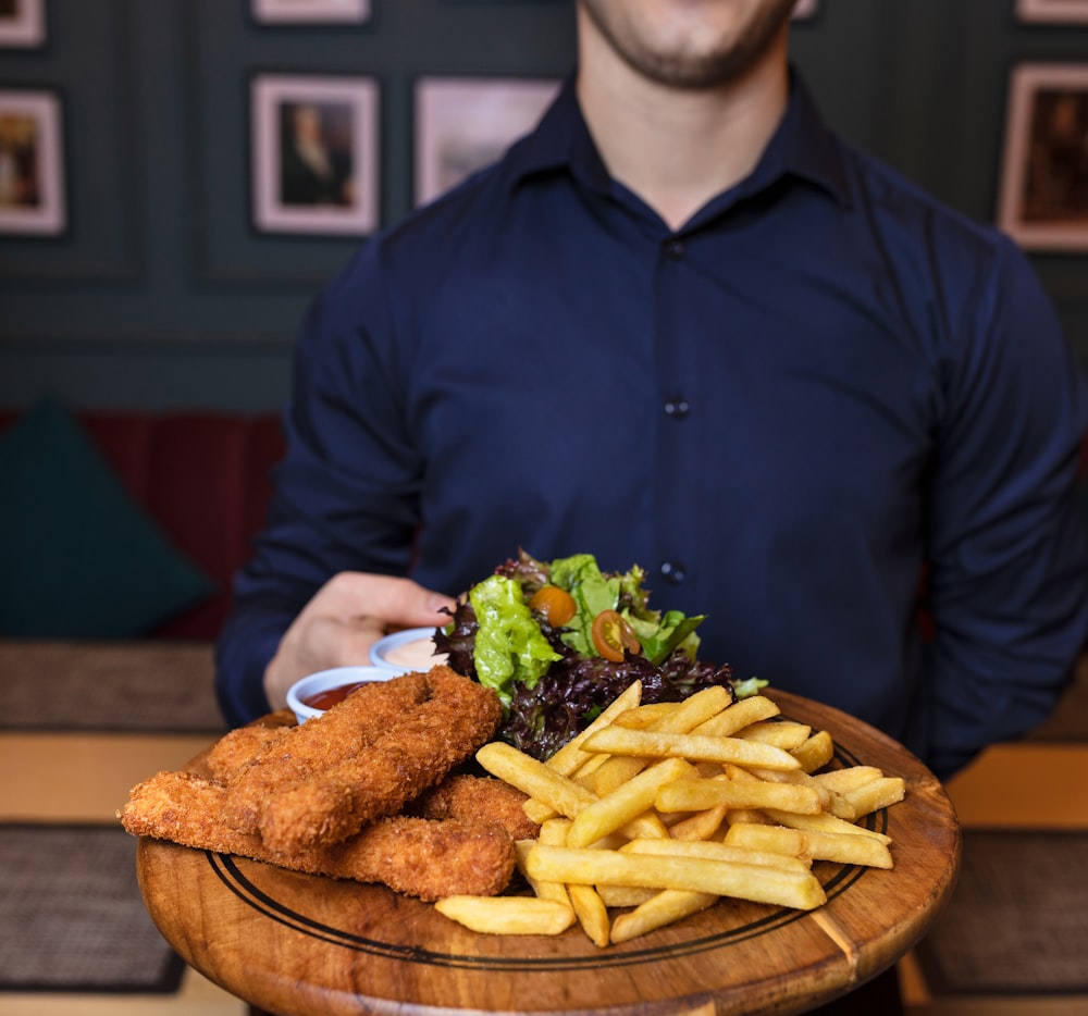 hombre con camisa de vestir azul sosteniendo una bandeja de madera marrón con papas fritas