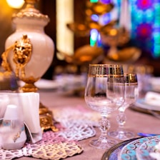 Elegant table setting featuring intricately decorated glassware with gold accents, ornate napkin holders, and a luxurious centerpiece with detailed designs. The background shows colorful stained glass patterns, enhancing the opulent atmosphere.