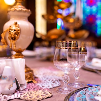 Elegant table setting featuring intricately decorated glassware with gold accents, ornate napkin holders, and a luxurious centerpiece with detailed designs. The background shows colorful stained glass patterns, enhancing the opulent atmosphere.