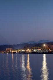 Twilight scene featuring a tranquil waterfront with reflections of bright lights from a distant shoreline. The lights illuminate palm trees and buildings along the coast, with mountains faintly visible in the background beneath a clear sky.
