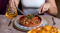 A person is dining with a fork and knife, enjoying a meal in a clay dish featuring a mix of vegetables and meat, possibly a stew. The table setting includes an intricately patterned plate beneath the dish, a wine glass with a golden liquid, and a piece of bread or pastry in the foreground.