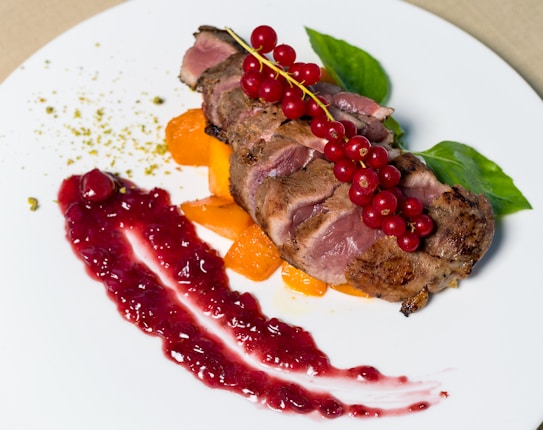 A gourmet dish featuring slices of seared meat topped with a cluster of red currants, garnished with vibrant green basil leaves. The plate is artfully arranged with a spread of red berry sauce, accompanied by cubes of orange vegetables, likely roasted sweet potatoes or carrots, and a sprinkle of green spices or herbs.