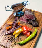 Grilled medium-rare steak slices are served on a wooden board alongside a colorful assortment of grilled vegetables, including red and yellow bell peppers, carrots, and grilled onions. A small green chili pepper adds a touch of spice. A sauceboat filled with brown sauce is placed at the top of the board.