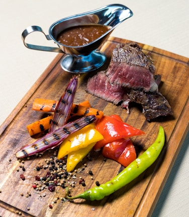 Grilled medium-rare steak slices are served on a wooden board alongside a colorful assortment of grilled vegetables, including red and yellow bell peppers, carrots, and grilled onions. A small green chili pepper adds a touch of spice. A sauceboat filled with brown sauce is placed at the top of the board.