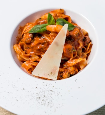 A plate of pasta topped with a rich tomato-based sauce, garnished with a slice of cheese and fresh basil leaves. The dish is elegantly presented on a white plate sprinkled with black pepper on the rim.