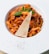 A plate of pasta topped with a rich tomato-based sauce, garnished with a slice of cheese and fresh basil leaves. The dish is elegantly presented on a white plate sprinkled with black pepper on the rim.