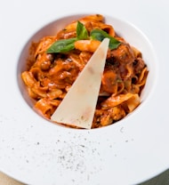 A plate of pasta topped with a rich tomato-based sauce, garnished with a slice of cheese and fresh basil leaves. The dish is elegantly presented on a white plate sprinkled with black pepper on the rim.