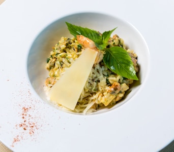 A gourmet dish featuring a creamy risotto garnished with a large triangle of shaved cheese and a sprig of fresh basil. The risotto includes visible pieces of shrimp and is served in a white bowl on a white plate, with a hint of red spice sprinkled on the side.