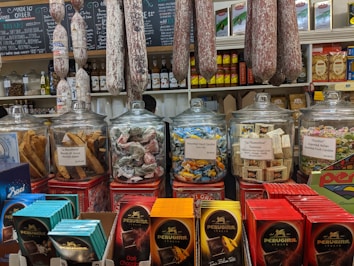 A variety of Italian food products are displayed in a deli-style setting. On the countertop, there are glass jars filled with different types of candies, biscotti, and Torrone. Labels indicate the contents, such as 'Assorted Hard Candies' and 'Puntini Imported Italian Assorted Fruit Candies'. Behind the jars, various packages of pasta and bottles of condiments are arranged on shelves. Large salami sausages are hanging above the countertop. The scene is colorful and inviting, showcasing a range of gourmet foods.