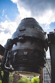 A large, cylindrical industrial structure made of black metal, featuring numerous bolts and rivets. The structure is positioned against a backdrop of a partly cloudy sky. Surrounding the structure are industrial elements and a glimpse of brick buildings and greenery.