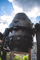A large, cylindrical industrial structure made of black metal, featuring numerous bolts and rivets. The structure is positioned against a backdrop of a partly cloudy sky. Surrounding the structure are industrial elements and a glimpse of brick buildings and greenery.