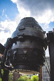 A large, cylindrical industrial structure made of black metal, featuring numerous bolts and rivets. The structure is positioned against a backdrop of a partly cloudy sky. Surrounding the structure are industrial elements and a glimpse of brick buildings and greenery.