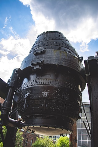 A large, cylindrical industrial structure made of black metal, featuring numerous bolts and rivets. The structure is positioned against a backdrop of a partly cloudy sky. Surrounding the structure are industrial elements and a glimpse of brick buildings and greenery.