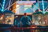 A person wearing a red cap and a blue shirt with text on the back is seen at a fairground or amusement park. They appear to be operating or managing one of the rides, with bright lights and colored fencing visible around the area. The scene is bustling with activity and has a lively atmosphere.