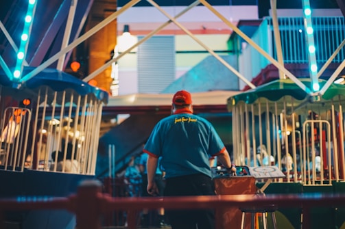 A person wearing a red cap and a blue shirt with text on the back is seen at a fairground or amusement park. They appear to be operating or managing one of the rides, with bright lights and colored fencing visible around the area. The scene is bustling with activity and has a lively atmosphere.